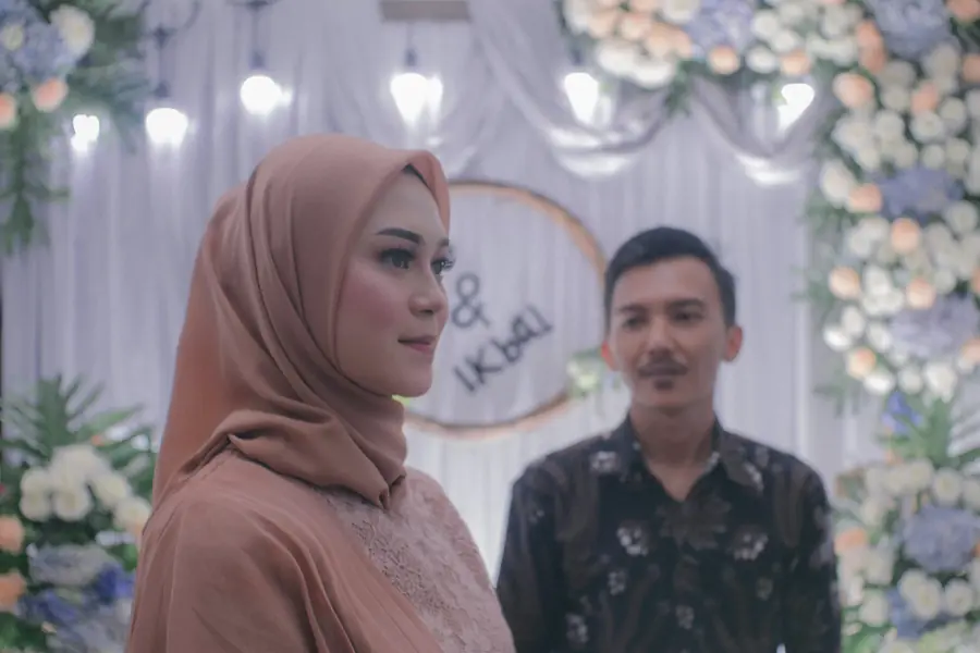 Pengantin berhijab merah muda berdiri di samping pasangannya di depan dekorasi bunga